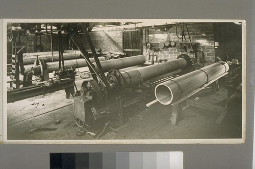 The pipe factory. Assembled pipe with temporary chain-clamps at right. Pipe in machine is being wound with heavy wire. Some pipes were later rolled in hot tar; then rolled in shavings