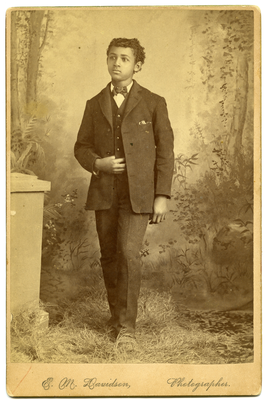 Unidentified boy wearing a dark suit with his right hand holding his vest