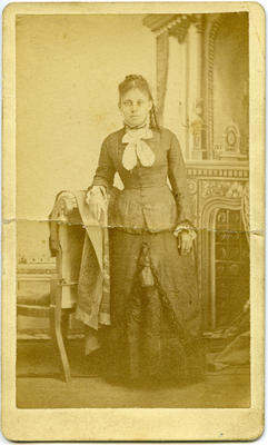 Woman standing wearing dark colored dress, right arm resting on arm of chair