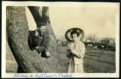 Man holding a top hat leaning against tree next to women holding a parasol