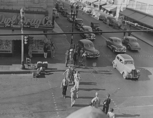 Pedestrians including men in uniform, cross W. Fourth Street in front of the Owl Drug Co