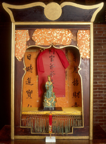 Wood shrine with figure of the God of Sun & Prosperity, 12" tall in wooden niche. Entire shrine placed on pine table 40" tall, painted dull red. Shrine painted a dull red with remnants of orange & gold temple money attached to outside. Inside of shrine covered with colored paper. Shrine figure is 12" tall & painted green & gold. May be ceramic