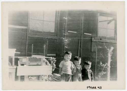 Photograph of three small boys standing in front of a bararcks with abandoned furniture