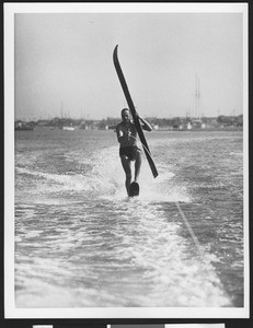 Man water skiing on a lake on one ski, holding the other in his hand, ca.1950