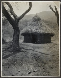 Person squatting in front of round house, Tanzania, ca.1926-1940