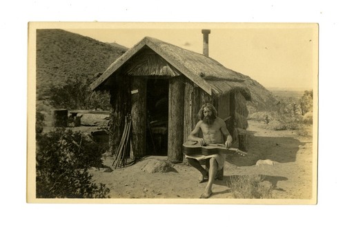 Bearded man with guitar in front of hut