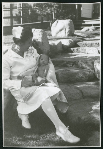 Photograph of Irene Gavigan, R.N., holding a newborn on the steps leading to the Manzanar hospital
