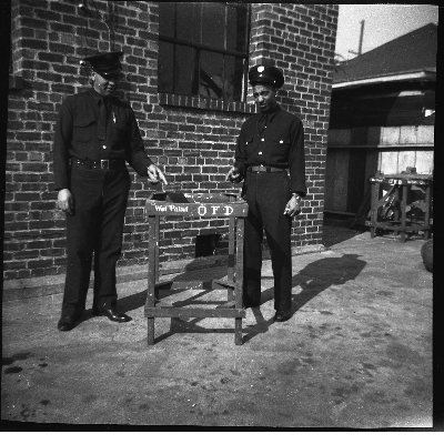 Two Oakland firefighters pointing to "Wet paint O.F.D." wood table