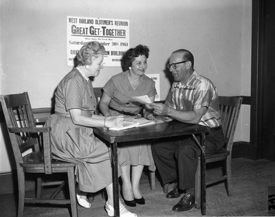 Two women and man seated at table with sign for West Oakland Oldtimer's Reunion