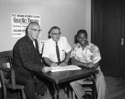 Three men seated at table with sign for West Oakland Oldtimer's Reunion