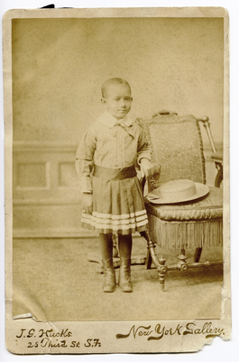 Portrait of unidentified child standing next to chair with straw hat