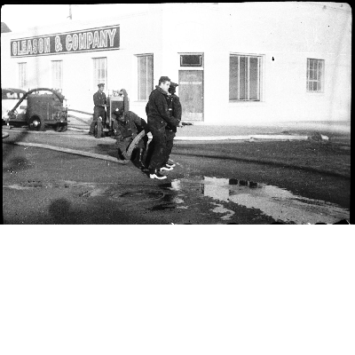 Firemen holding fire hose in street next to Gleason and Company building