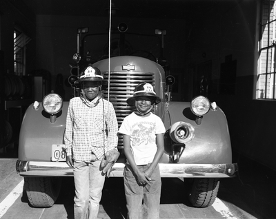 Two boys wearing fire helmets standing in front of fire truck