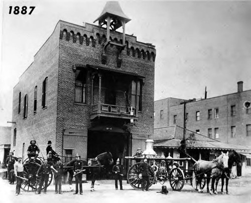 Photograph of a group of firemen standing in front of the Firehouse, built in 1884 just south of the Plaza, with an Ahrens steamer