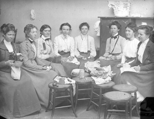 [Group portrait of eight women eating food in a classroom]