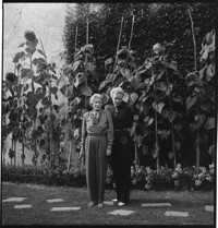 [Janet Flanner in garden with unidentified woman, both wearing pantsuits]