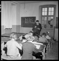[First French Lesson: small child, Hans Weibel, in dark (Alsatian?) suit and round hat, in school room with older children]