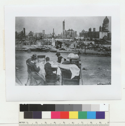 [Refugees eating at table in street adjacent to ruins. City Hall in distance, right.]