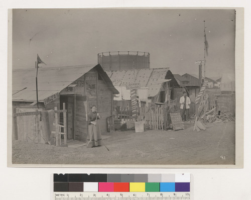 Refugees living in houses made from dry goods boxes. S.F. Cal. Near Lobos Square. Refugees of S.F. Cal. 1906. These houses are made from boxes. April 30, 1906. [verso:] Immediately after the fire. [Woman with broom and man standing before refugee shacks. Unidentified location.]