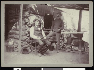 Two fishing club members outside a mountain residence, showing a kitchen area, ca.1910-1920