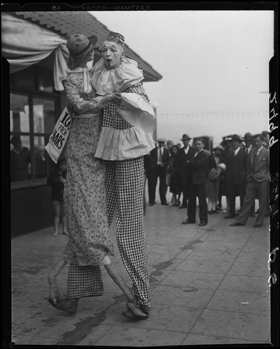 Clown on stilts dancing with tall puppet, advertising dance marathon, Santa Monica, 1928