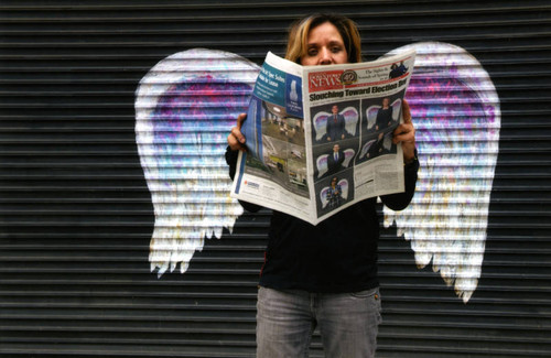 Unidentified woman holding a newspaper posing in front of a mural depicting angel wings