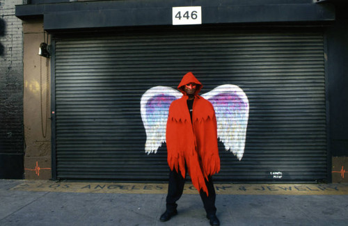 Unidentified person in a red hooded cape and mask posing in front of a mural depicting angel wings