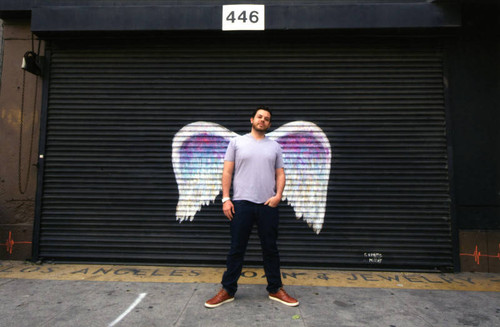 Unidentified man in a grey shirt posing in front of a mural depicting angel wings