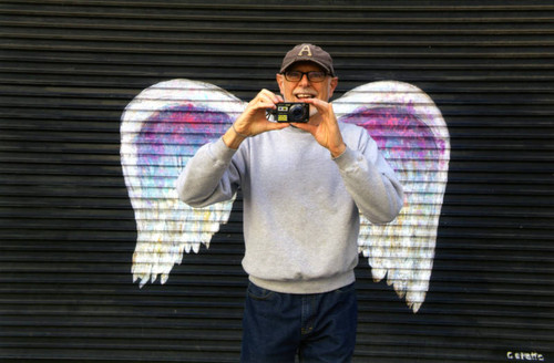 Unidentified man holding a camera posing in front of a mural depicting angel wings