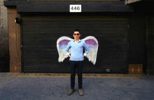 Unidentified man in sunglasses and light blue sweater posing in front of a mural depicting angel wings