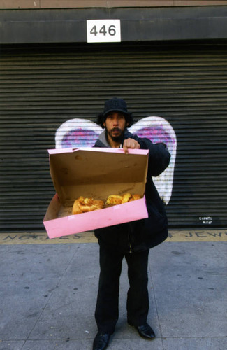 Unidentified man holding an open pastry box posing in front of a mural depicting angel wings