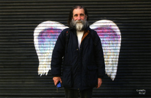 Unidentified man with facial hair posing in front of a mural depicting angel wings