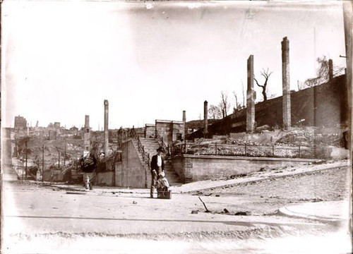 [Unidentified child and one other person standing in front of decimated buildings]