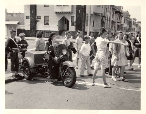 [Patrolwoman sits on her three wheeled motorcycle while two girls help young people cross the street]