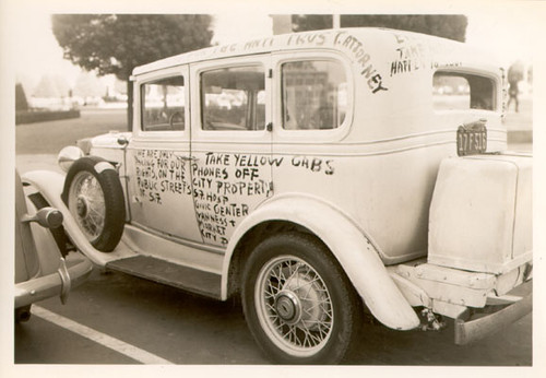 [Taxicab with protest messages written on it parked across from City Hall]