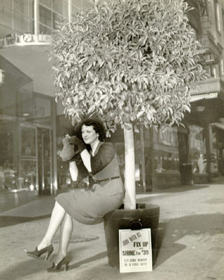 [Woman looking into a compact mirror sitting on tree planter box next to a sign that reads 'Join With Us Fix Up Shine for '39']