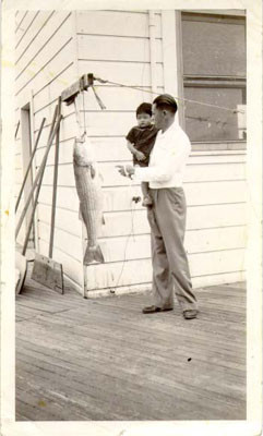 [Unidentified man with a small child and a large fish on a porch in Visitacion Valley]