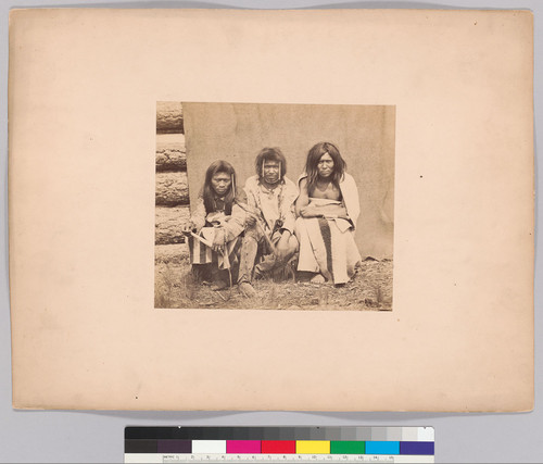 [Three Kootenai men, full-length portrait, seated against blanket in front of a log cabin, facing front, wearing traditional dress.]