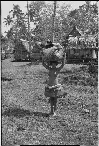 Young woman in short fiber skirt holds large basket on her head, village houses in background