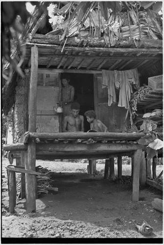 Family scene: man and child with a book, woman with tea kettle in doorway of small house