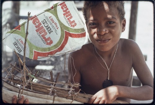 Model canoe with sail made of rice bag, held by smiling boy in Wawela village