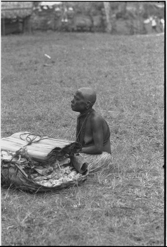 Mortuary ceremony, Omarakana: mourning woman, face and body covered with ash, with basket of banana leaf bundles and mats