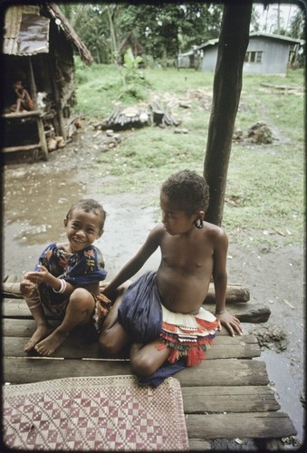 Children sitting on house veranda next to woven mat, girl (r) wears short fiber skirt and turtle-shell earrings