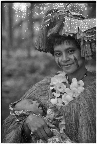 New mother, holding her infant, wears long fiber shawl, flower garland, and skirt valuable on head, paint on face