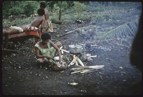 Food preparation: Kitemwalusa peels yams for cooking, girl in red skirt behind her