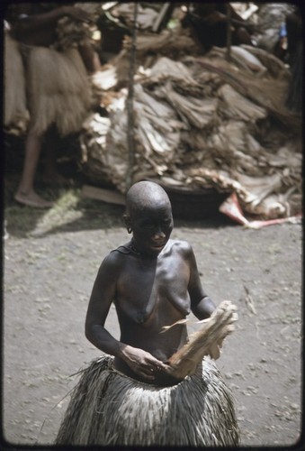 Mortuary ceremony: mourning woman, ash-blackened skin and shaved head, presents banana leaf bundle at exchange, large pile of bundles in background