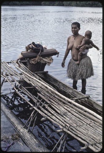 Canoes: woman holds infant, near a canoe used for coastal transport with basket and other items on board