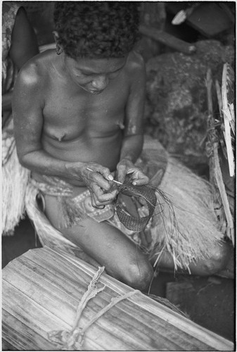 Weaving: woman weaves armband, she wears decorated fiber skirt, rolled matt in foreground