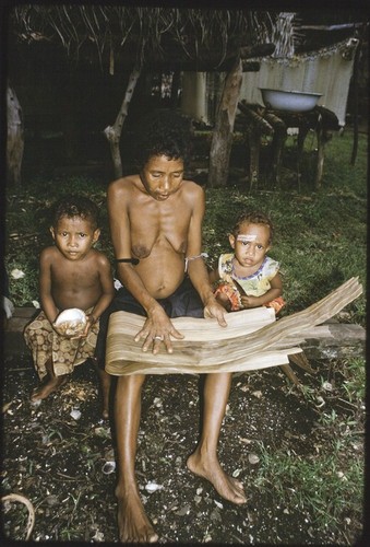 Woman folds strips of dried pandanus leaves (to be woven later), two children at her side