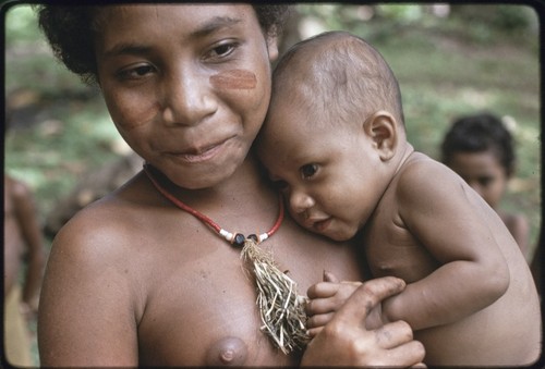 Adolescent girl, wearing shell necklace and dabs of betel nut paste on her cheeks, cuddles a baby and smiles